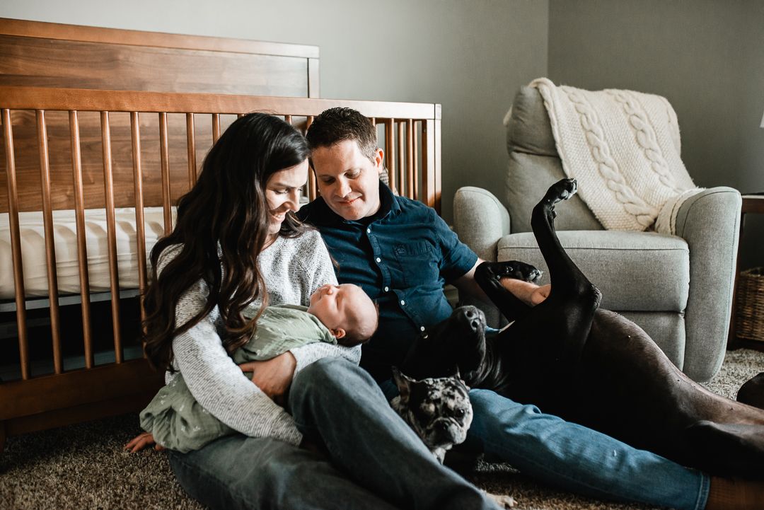 Family with newborn baby sits on nursery floor beside crib with dog.