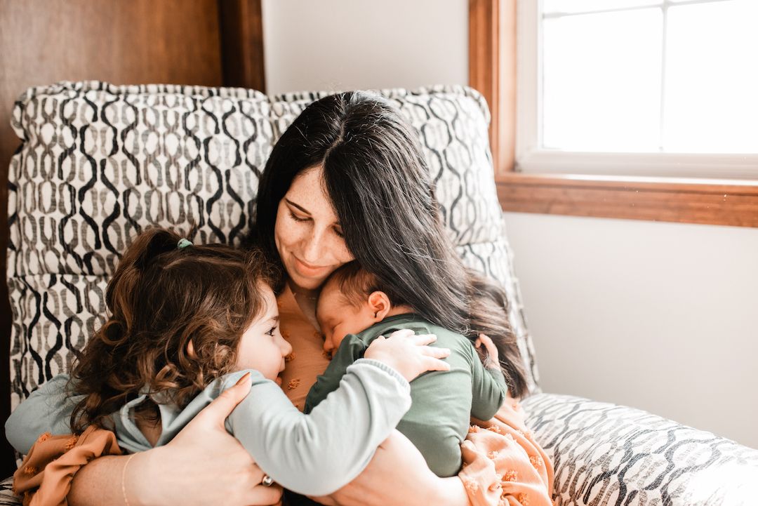 A mother with dark hair tenderly embraces her toddler and newborn baby while sitting in a cozy chair by a window.