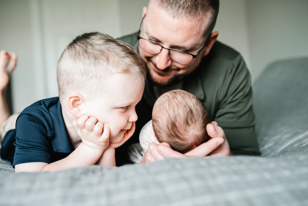 Smiling father with glasses introducing newborn baby to curious toddler son on bed.