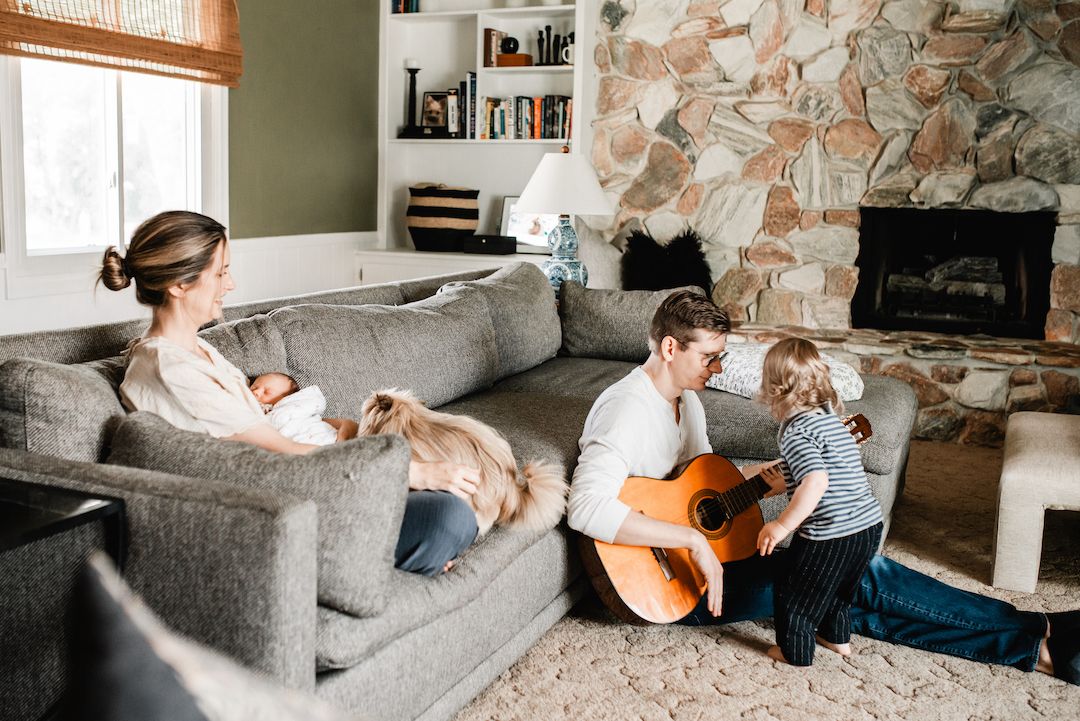 Family relaxing in cozy living room, mother holding newborn on sofa while father plays guitar for toddler.