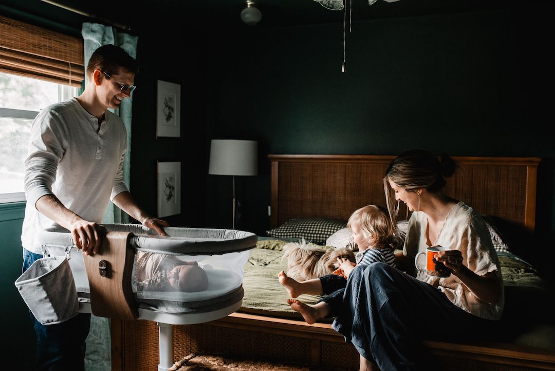 Young family in bedroom, father tending to baby bassinet while mother sits on bed with toddler in warm home setting.