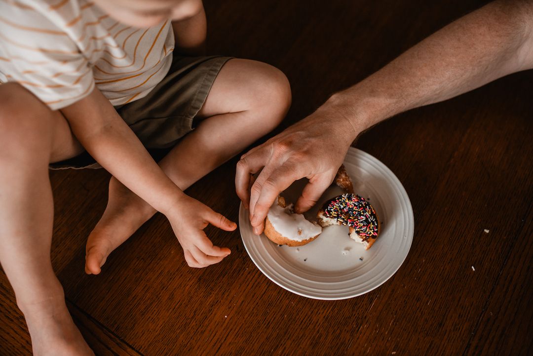 Adult and child sharing a sprinkle donut on wooden floor together.