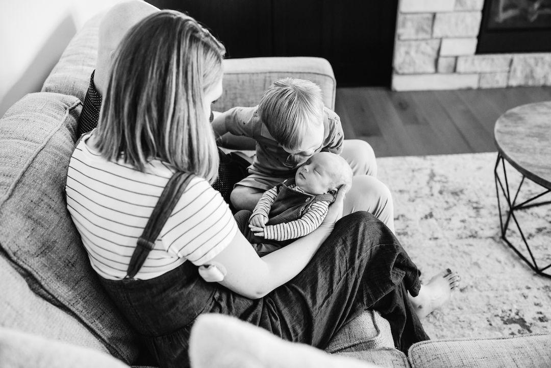 Mother holding newborn while young child leans in for a closer look on living room sofa.