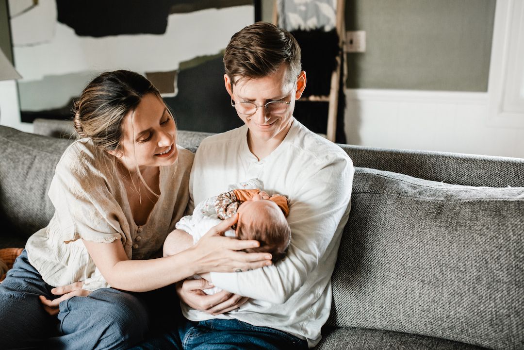 Young couple sitting on gray sofa admiring their newborn baby together in a cozy living room.