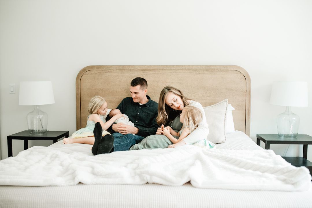 Family of four cuddles together on a white bed, with parents embracing two young children in a bright, cozy bedroom.