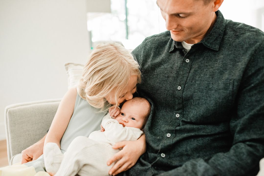 Father sitting with toddler and newborn baby, sharing a tender family moment in a bright, cozy living room.