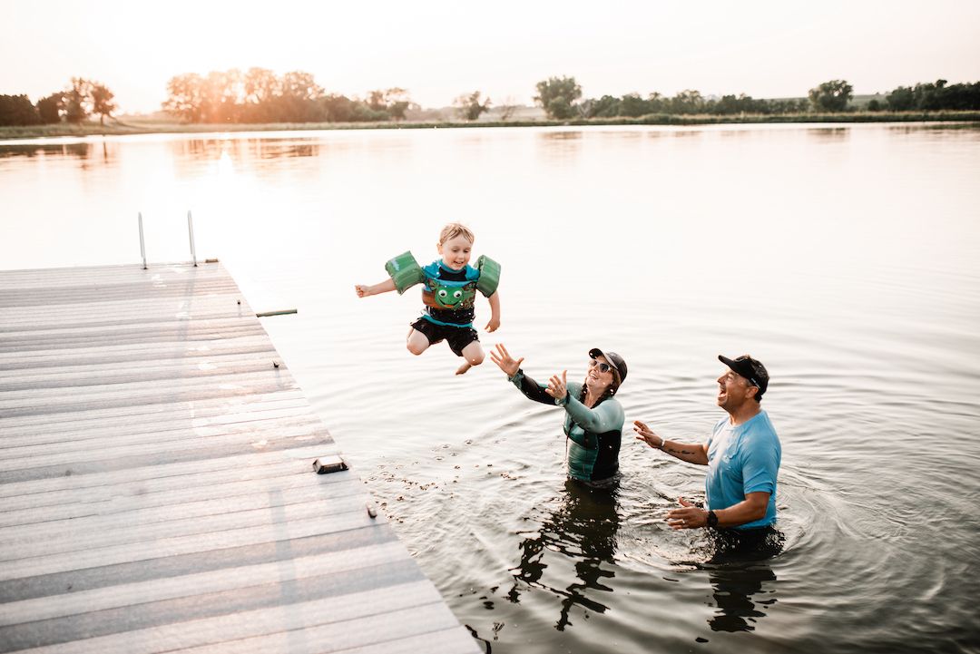Two men in a lake catching a young boy jumping off a wooden dock at sunset.