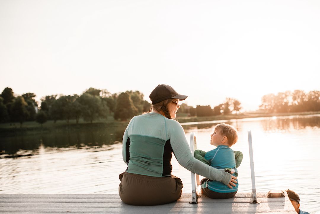 Mother and young child sitting together on wooden dock watching sunset over calm lake.