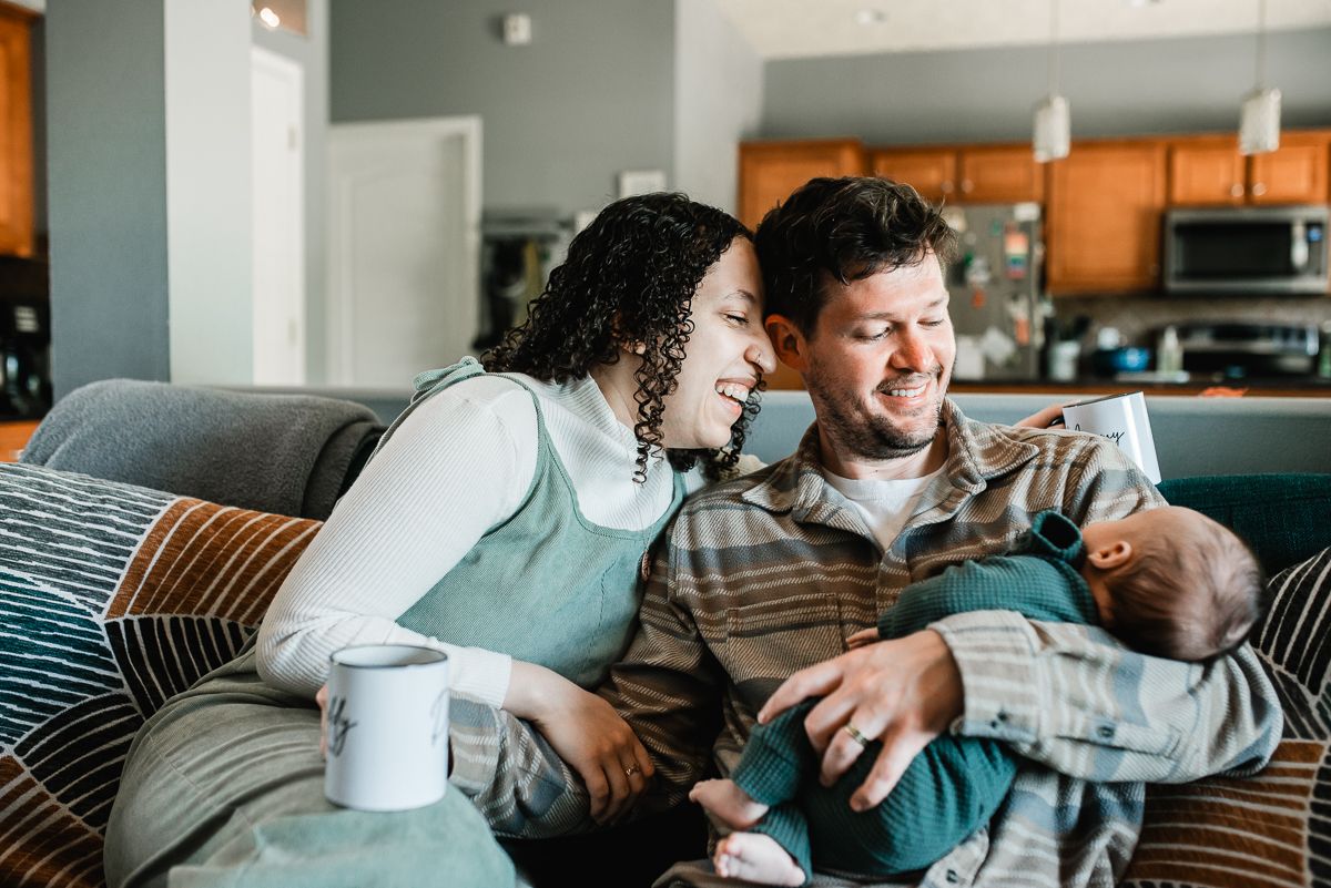 Happy couple sitting on couch smiling at newborn baby in cozy apartment living room.