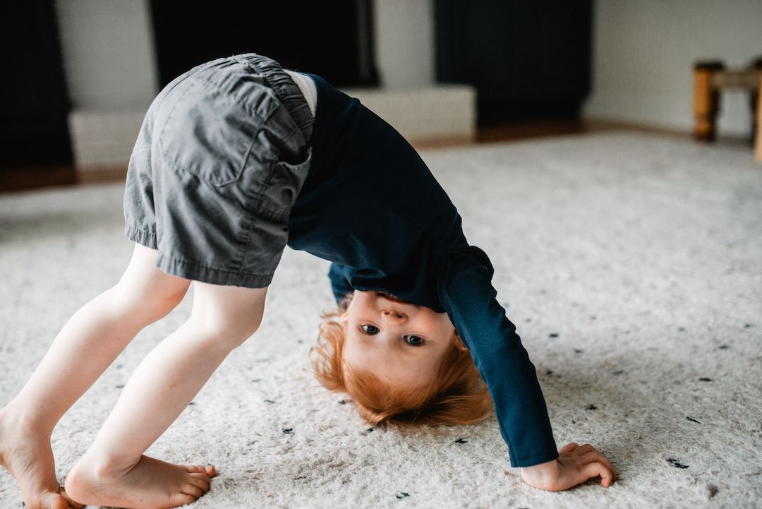 A young toddler does a backbend on a carpet indoors, playing and exploring movement at home.