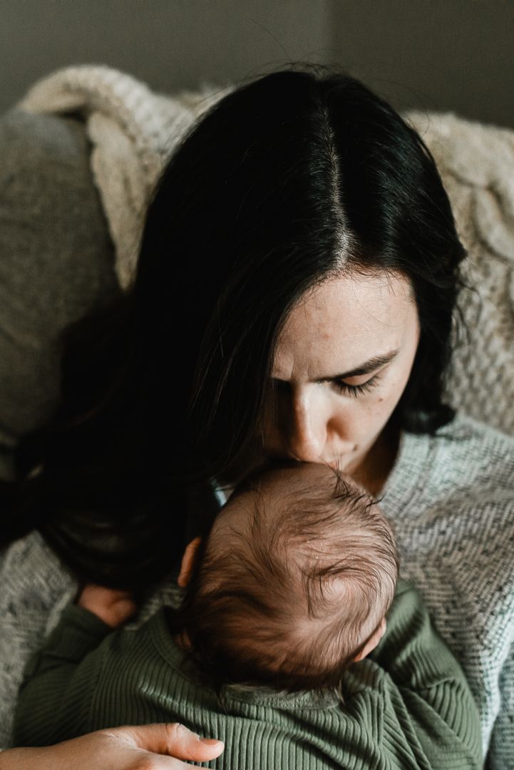 A dark-haired mother tenderly kissing her newborn baby's head while cradling them gently.