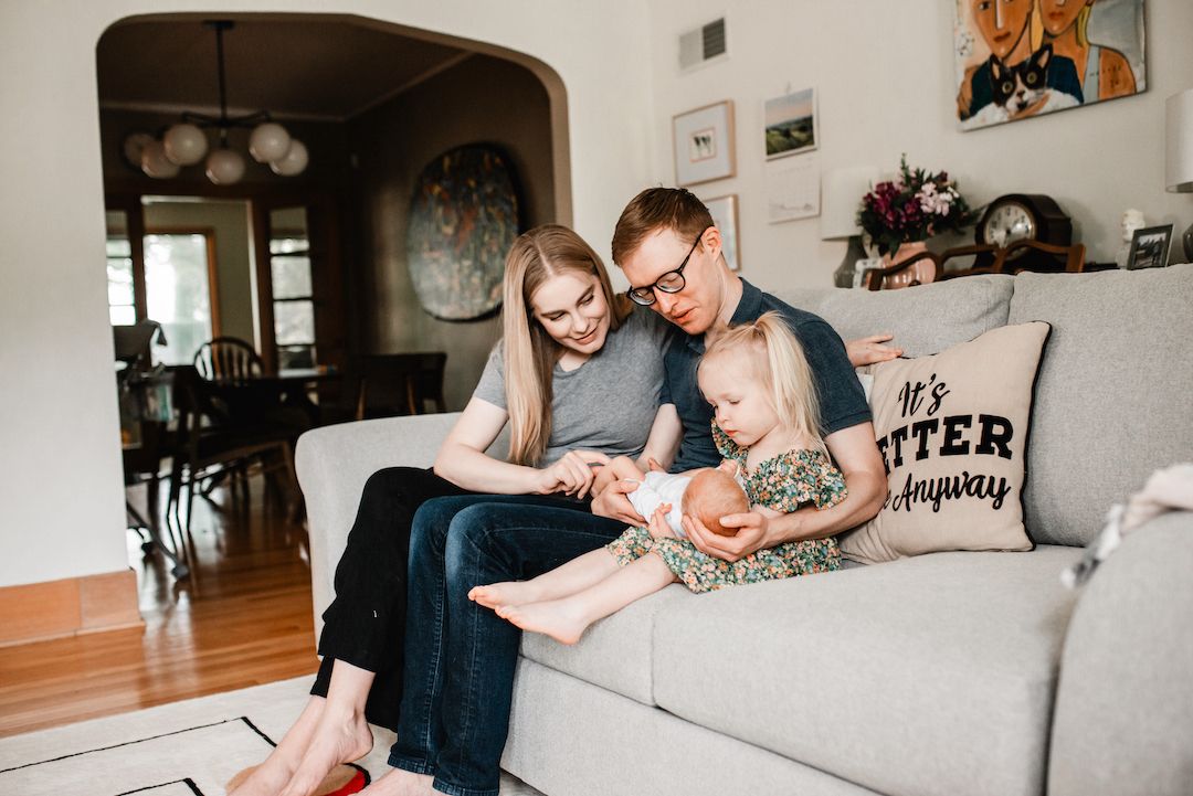 A young family sits together on a gray couch in a cozy living room, parents smiling at their toddler daughter.