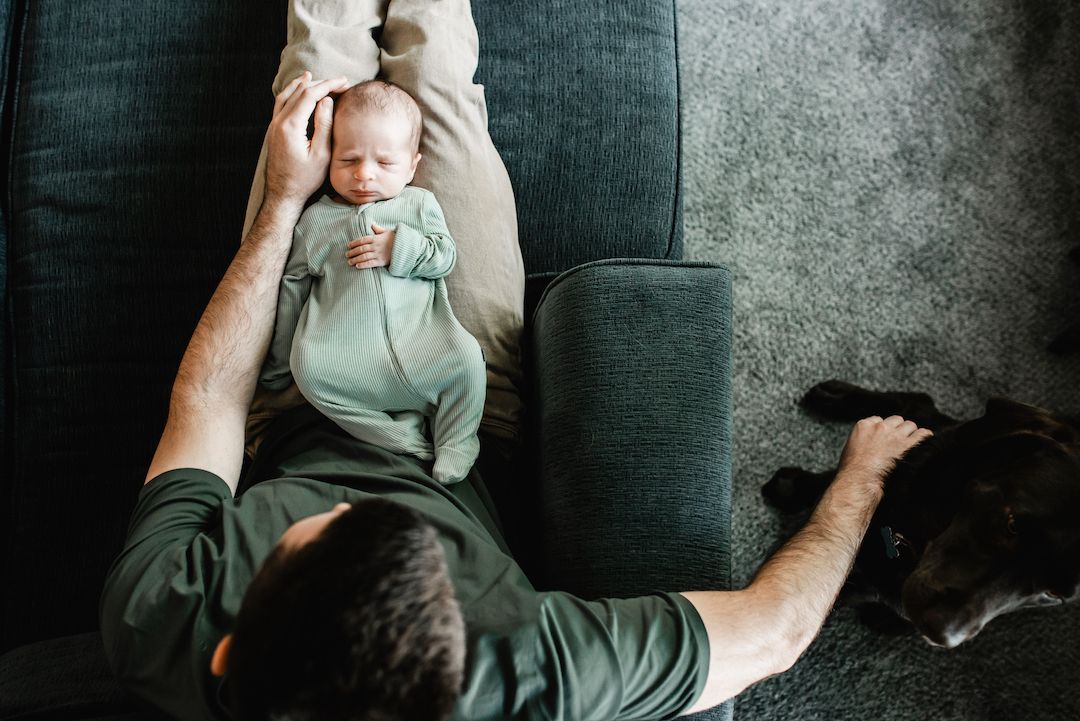 Overhead view of a father lying on a sofa holding his newborn baby in a mint green onesie.