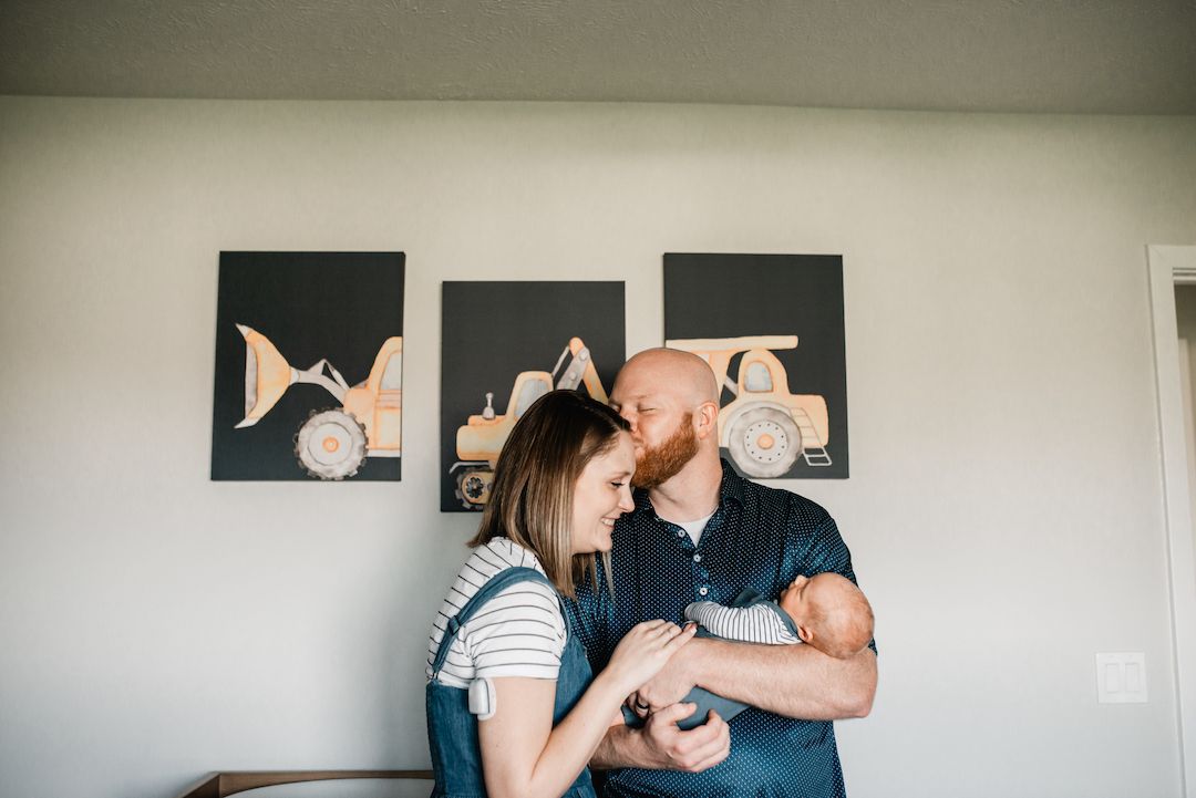 A couple shares a tender kiss while holding their newborn baby in a nursery with construction vehicle wall art.