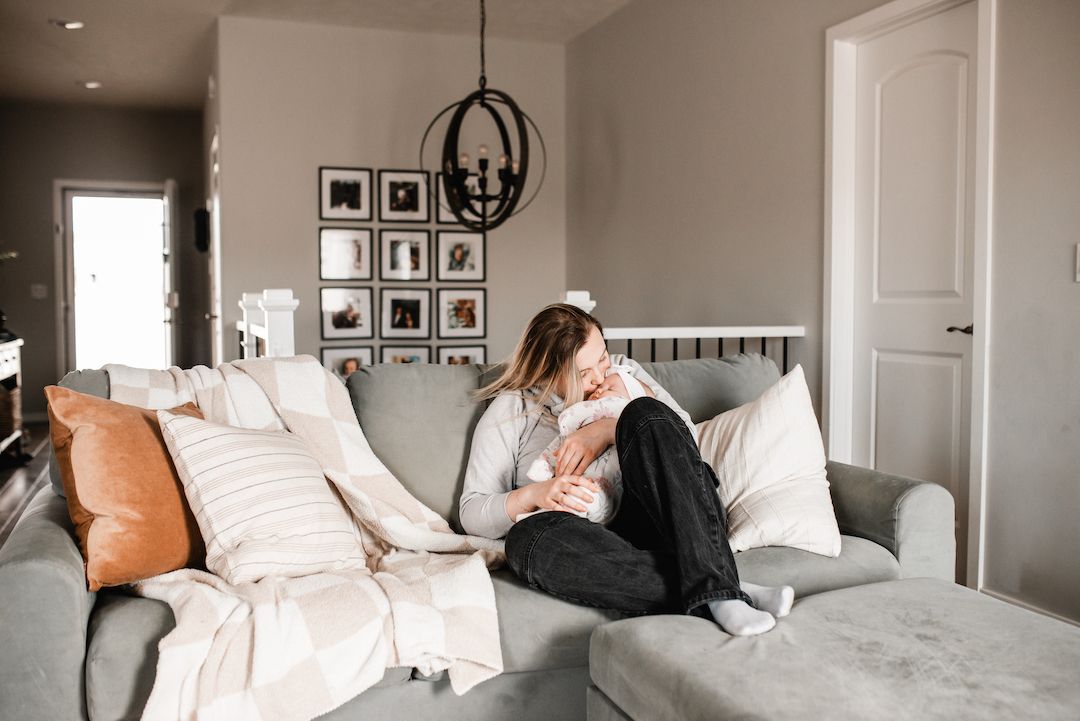 A mother tenderly holds her newborn baby while sitting on a cozy gray sofa in a modern living room.