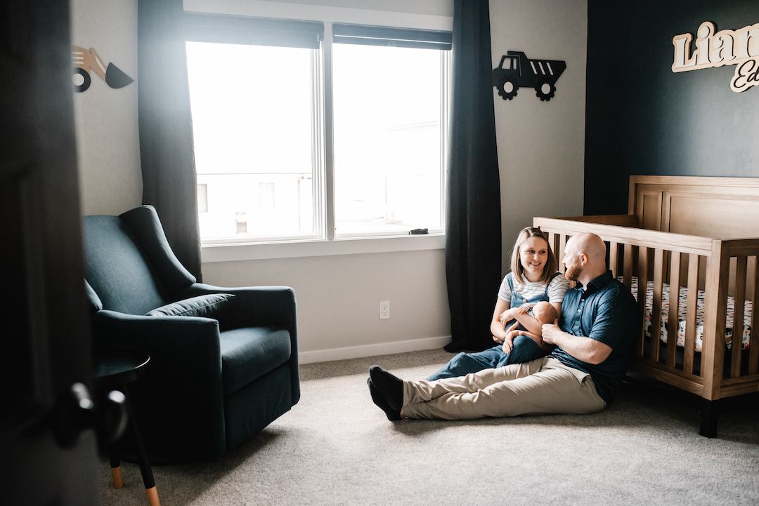 Parents sitting on nursery floor with baby near wooden crib in dark-themed boy's room with truck decor.