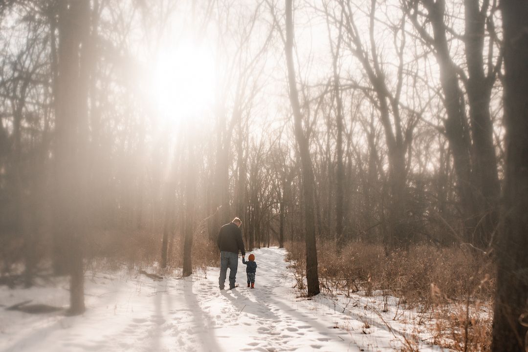Parent and child walking hand in hand through a snowy sunlit forest path.