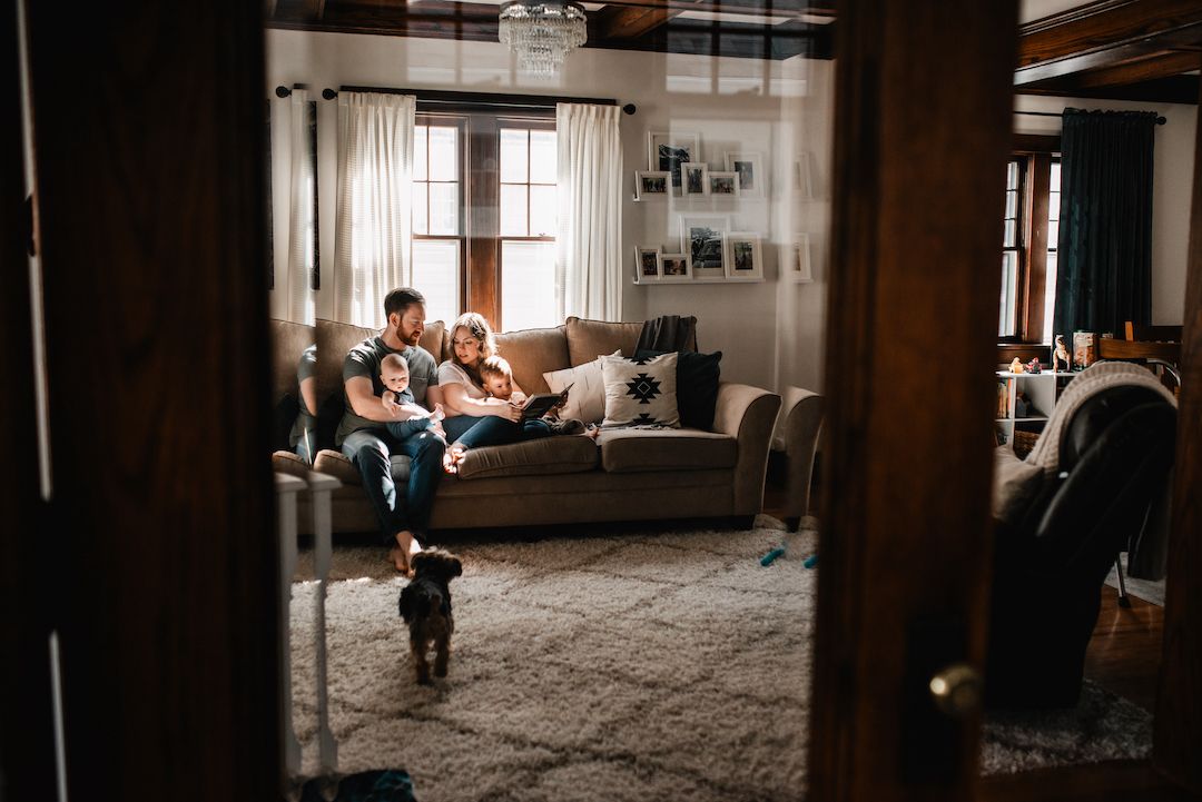 Couple relaxing on couch with small black dog playing on rug in cozy living room viewed from doorway.