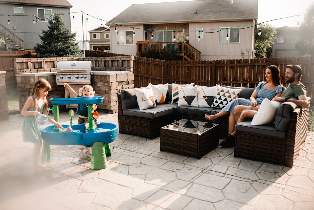 Family relaxing on patio furniture while children play at a water table in a backyard.