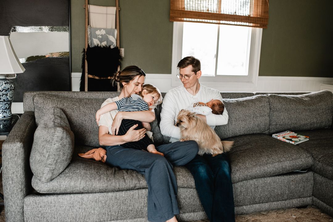 Young family with baby and small dog relaxing together on gray couch in cozy living room.