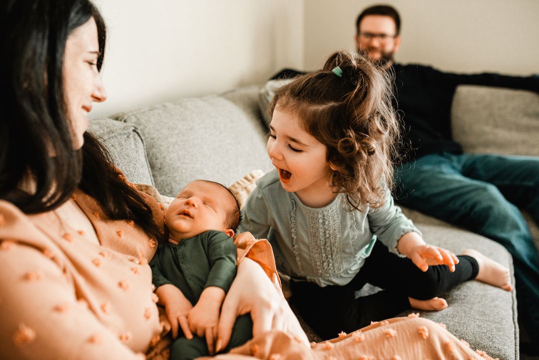 Excited young girl reacts to newborn sibling held by mother on couch with father behind.