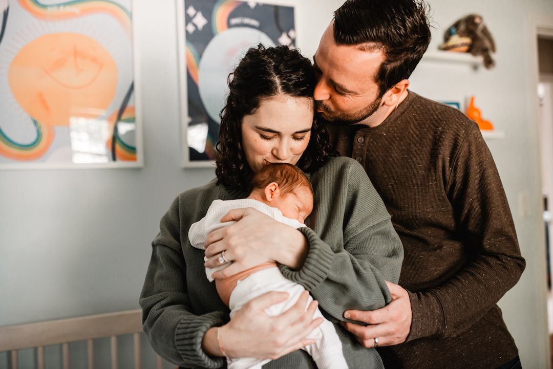 New parents tenderly holding their newborn baby in a cozy nursery room with colorful wall art.