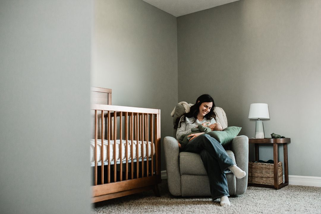 Mother sitting in nursery glider chair holding newborn baby next to wooden crib in soft gray room.