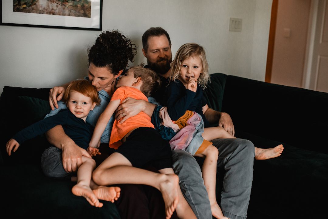 Two parents cuddle with their three young children on a dark couch indoors.