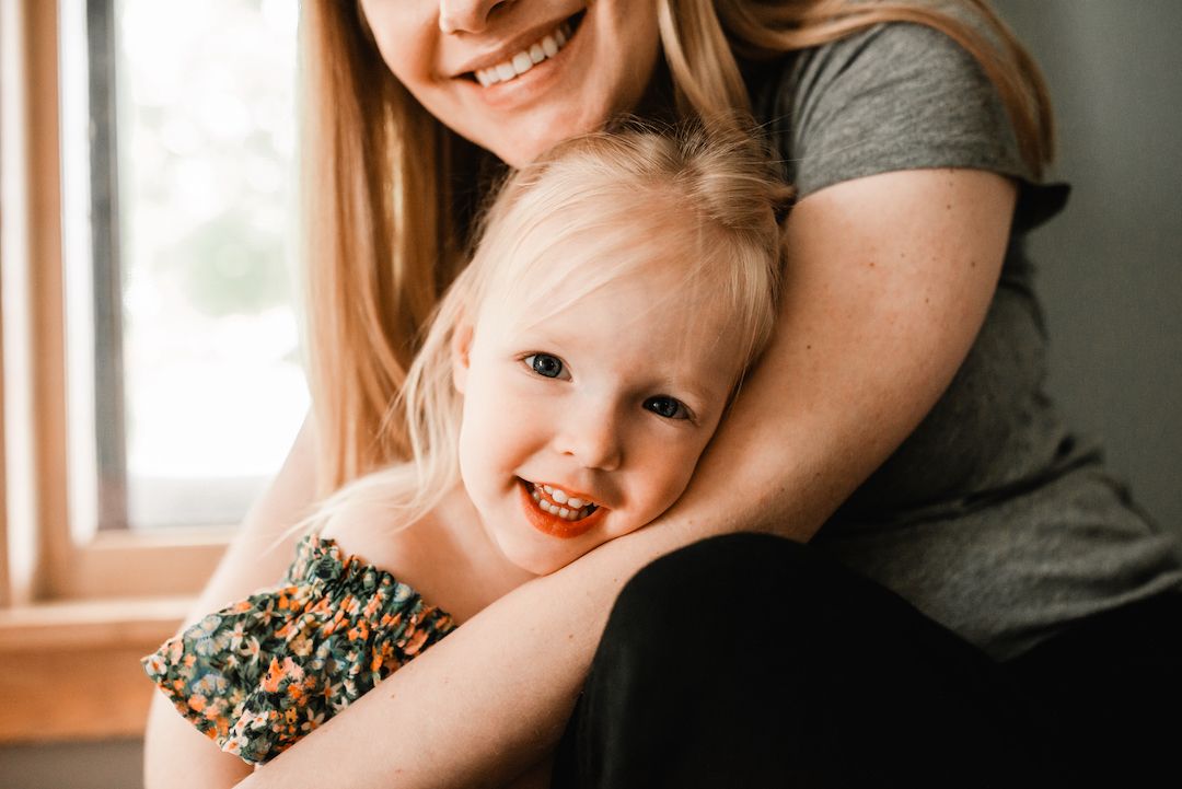 Mother hugging smiling blonde toddler girl by a bright window in a lifestyle portrait.