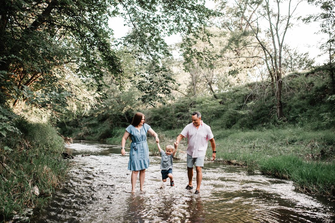 Young family holding hands and walking through a shallow creek surrounded by lush green trees.