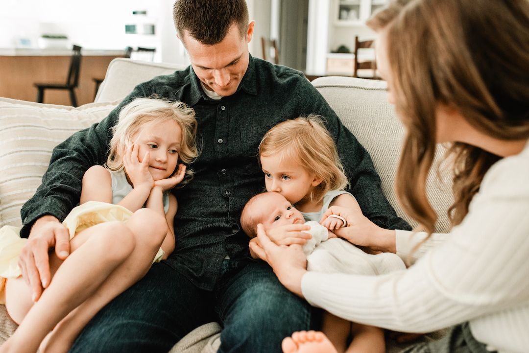Family of five sitting on couch, father holding newborn baby with two young daughters smiling beside him.
