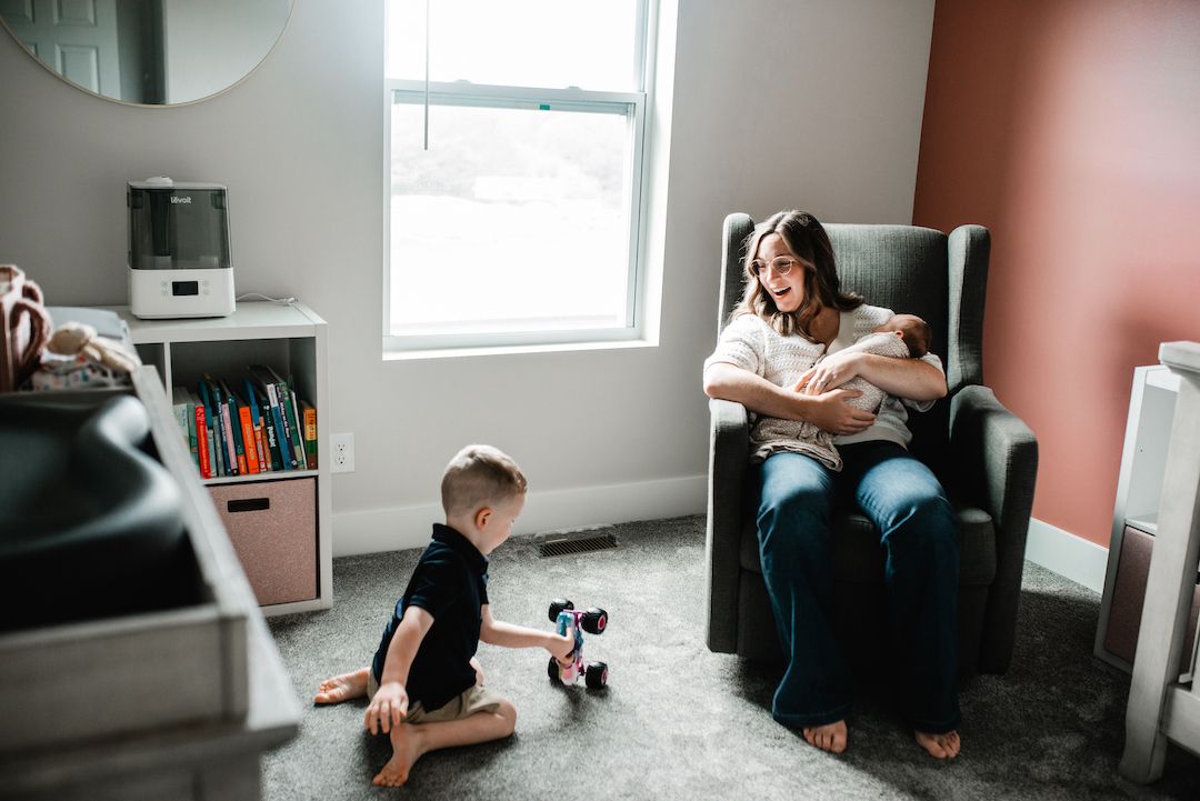 Mother sits in gray glider holding newborn while toddler plays with toy on floor in nursery.