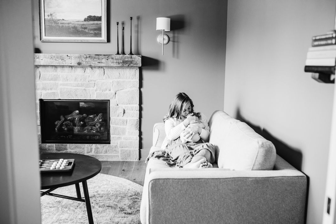 Black and white photo of a mother cradling her newborn on a cozy sofa beside a stone fireplace.