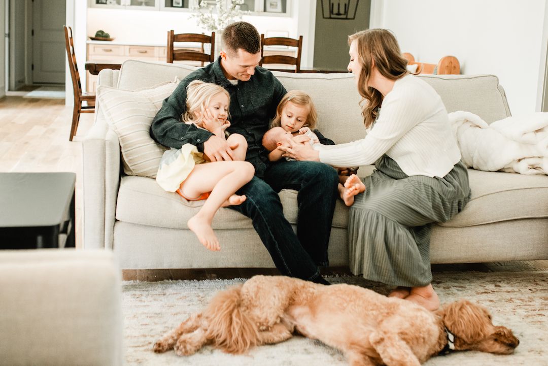 Family of four sitting on a cozy couch with a golden retriever dog resting on the floor in a bright living room.