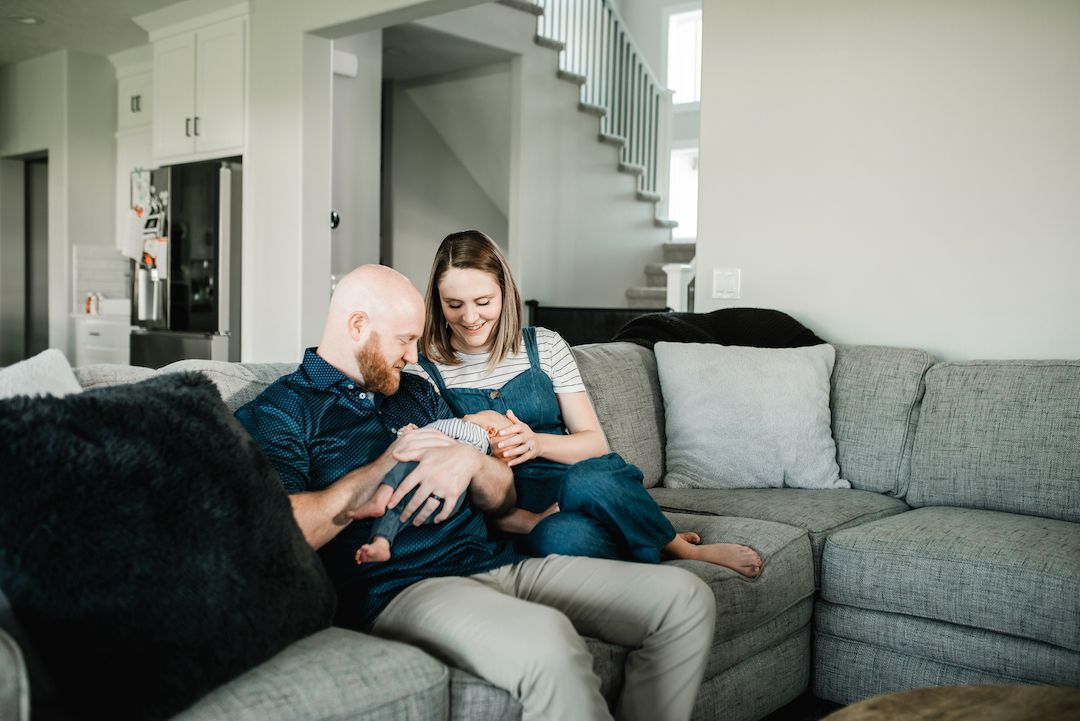 Happy couple sitting on a gray couch together admiring their newborn baby in a modern home.