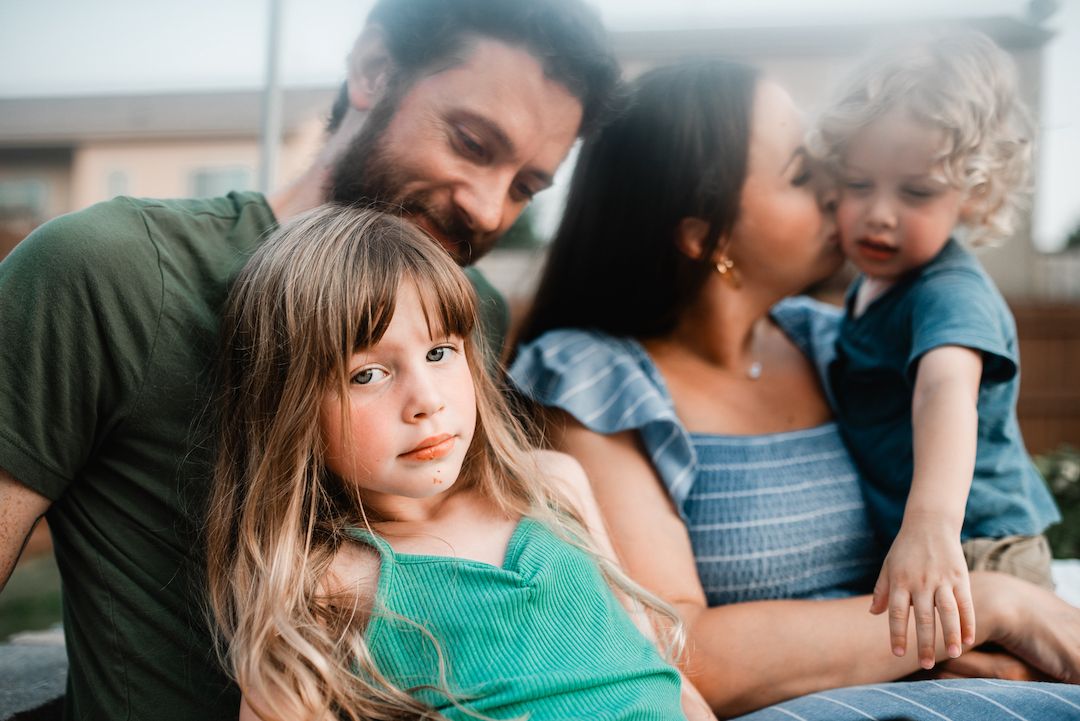 Close-up portrait of a family with two young children sitting together outdoors at dusk.