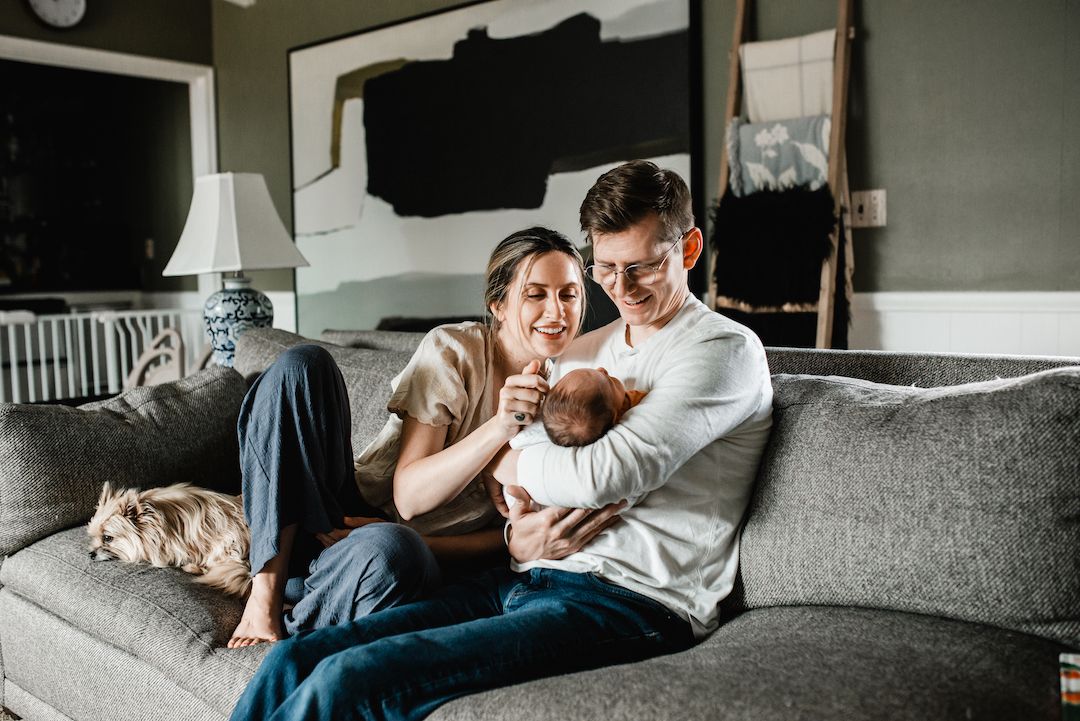 Happy couple sitting on gray sofa with newborn baby and dog in cozy living room.