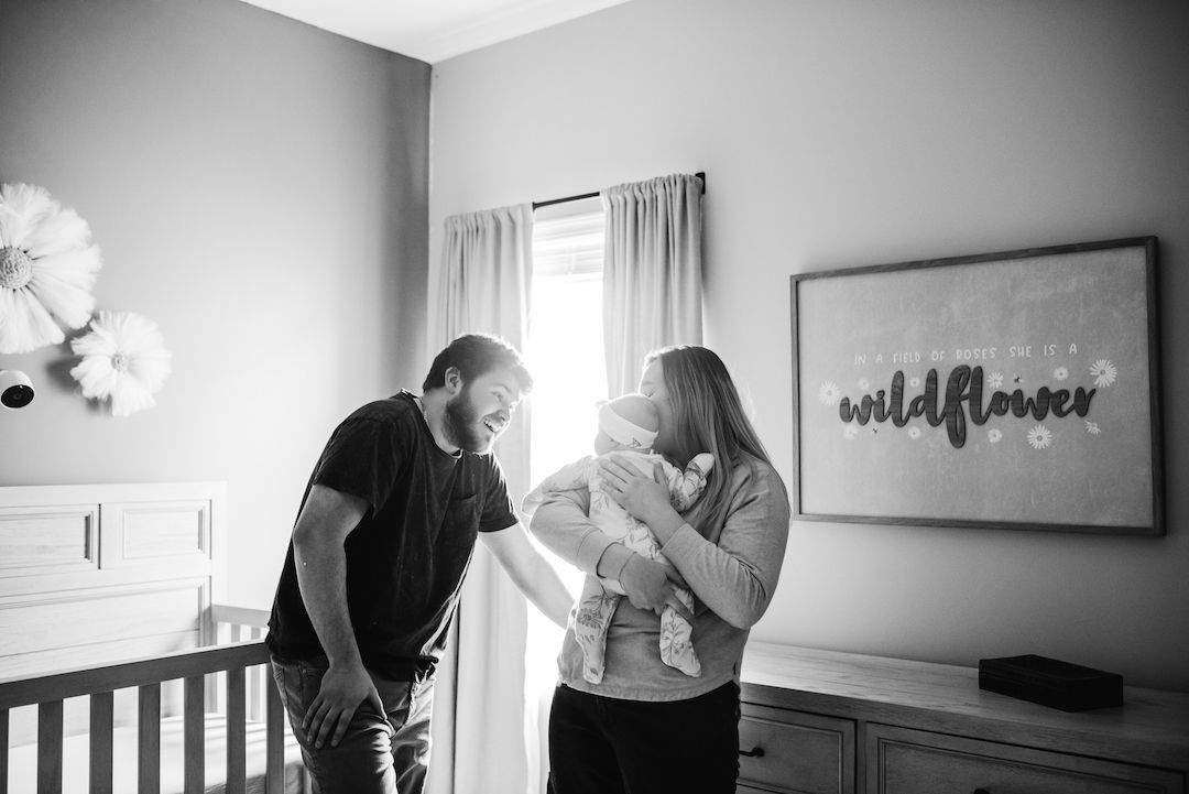 Black and white newborn photo of parents admiring their baby in a nursery with a wildflower sign.