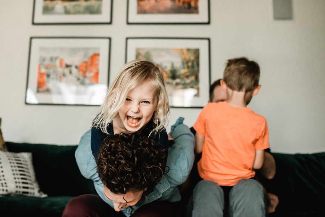 Laughing blonde girl being carried on back of adult while boy sits on dark couch behind them.