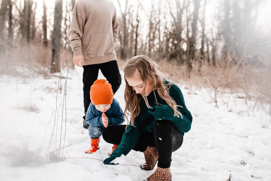 Older sister helping toddler in orange hat explore snow in a winter forest.