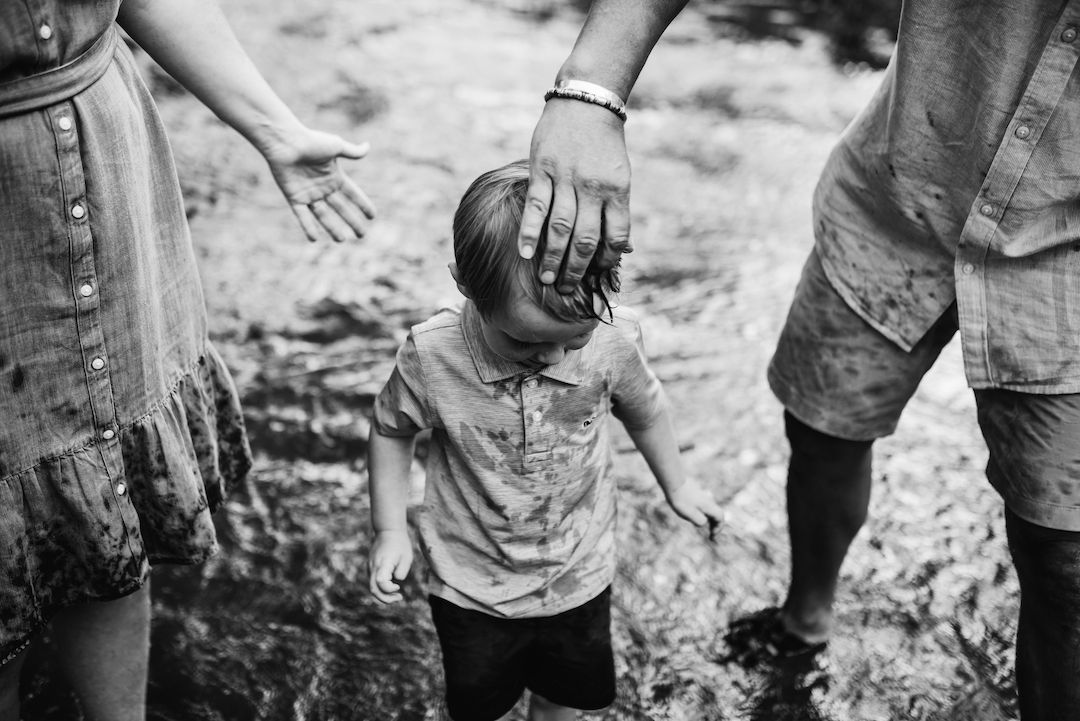 Black and white photo of parents holding hands with a young toddler outdoors.