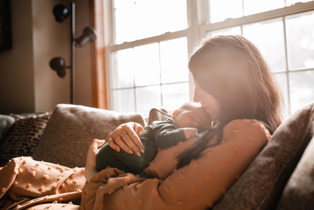 Mother tenderly breastfeeding newborn baby on couch in warm sunlit living room.