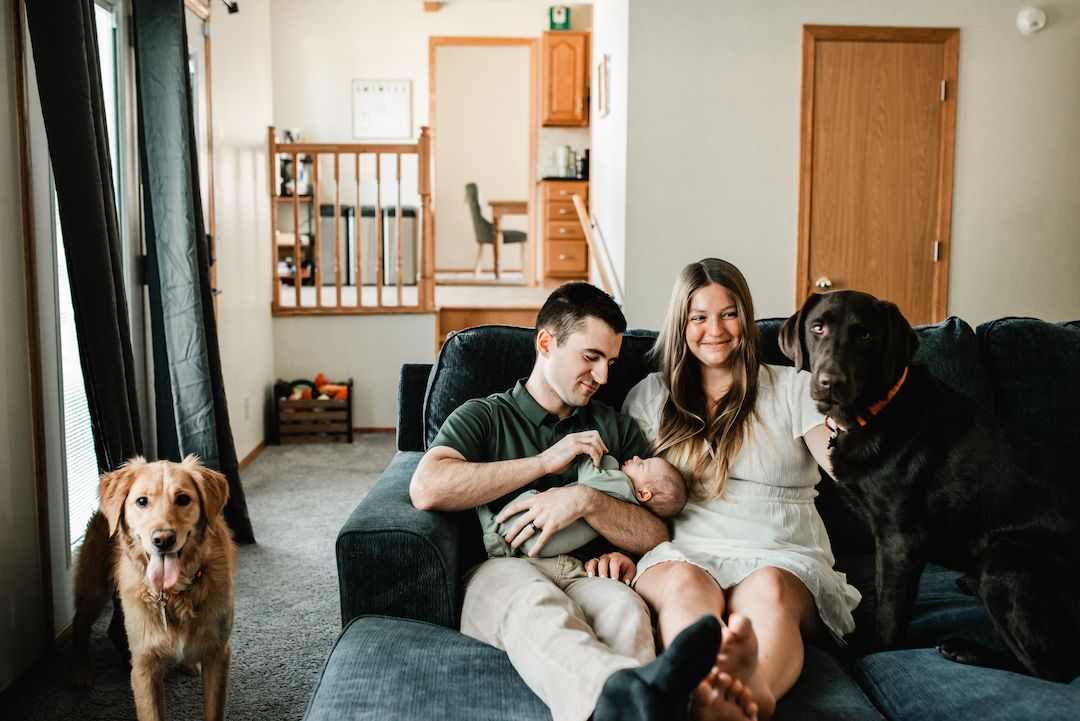 A couple sits on a gray sofa with their newborn baby and two dogs in a cozy living room.