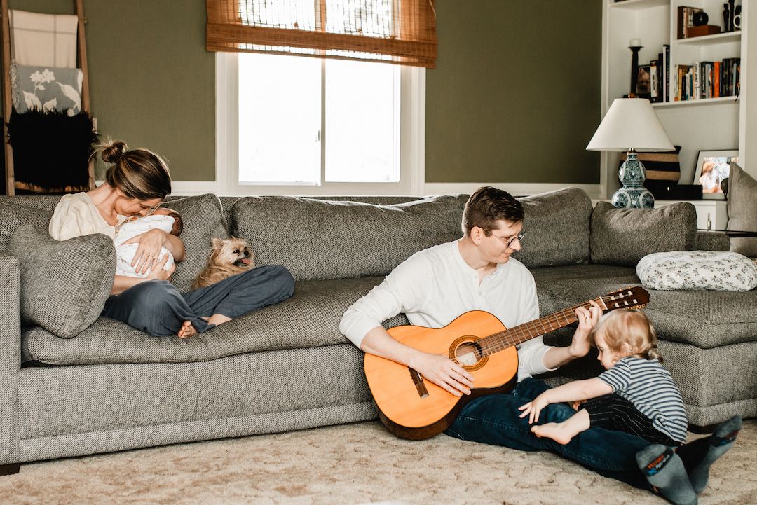 Family relaxing at home, father playing acoustic guitar while mother holds newborn baby on couch.