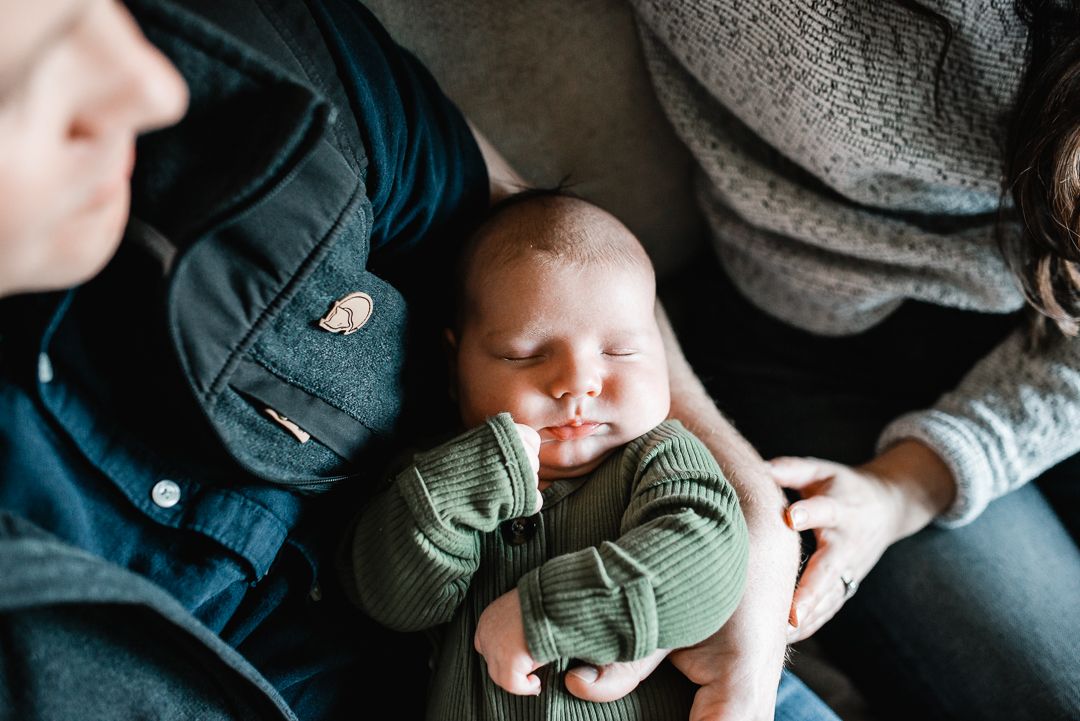 Sleeping newborn baby in green onesie cradled between two parents on a couch.