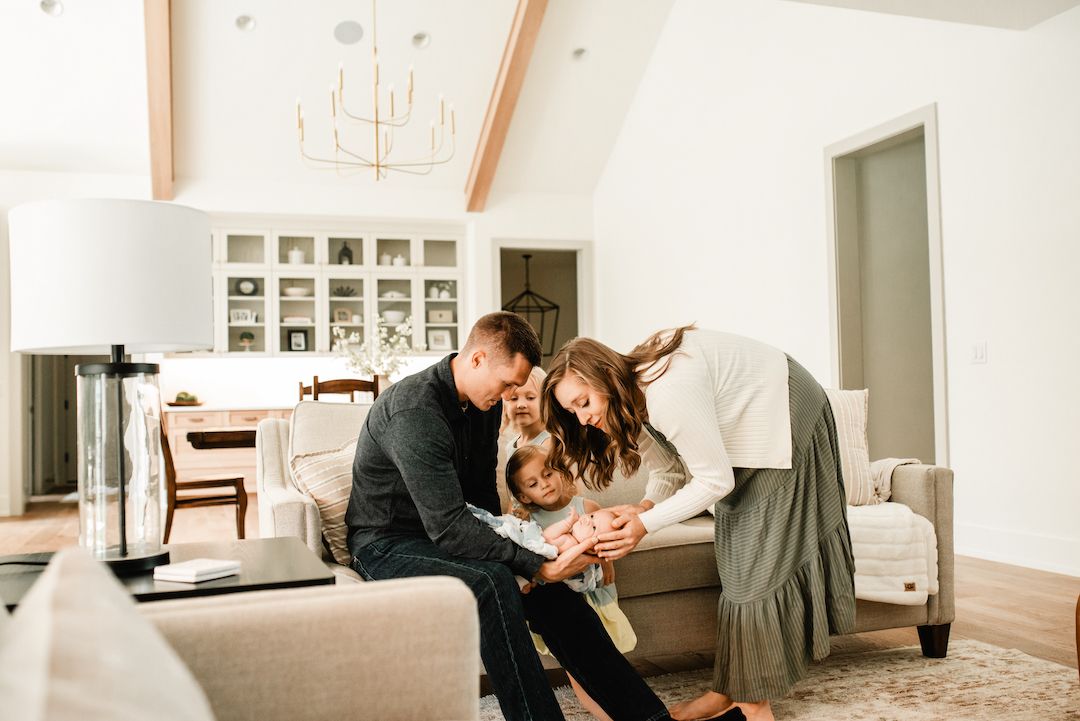 Happy family of four huddled together on sofa in bright modern living room, admiring their newborn baby.