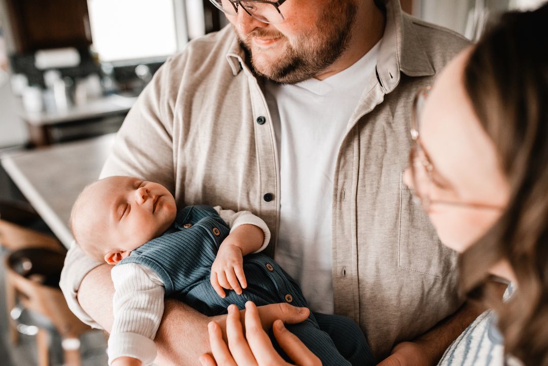 A father holds his sleeping newborn baby while a woman looks on adoringly in a warm home setting.