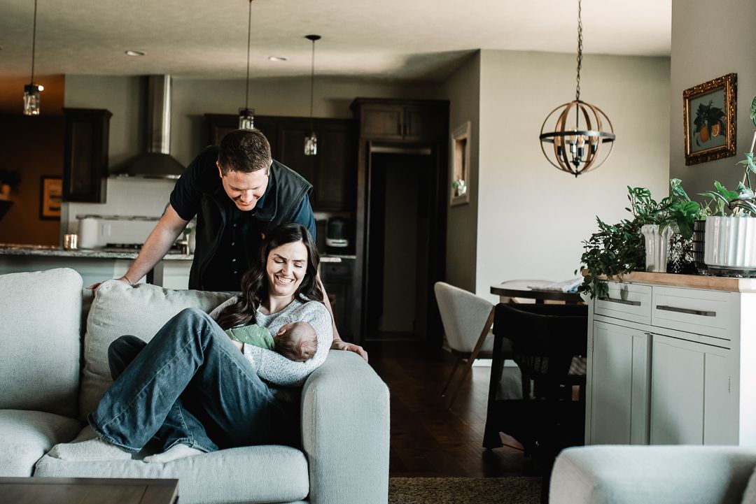 Happy couple relaxing on gray sofa in modern living room, woman holding newborn baby while man leans over smiling.