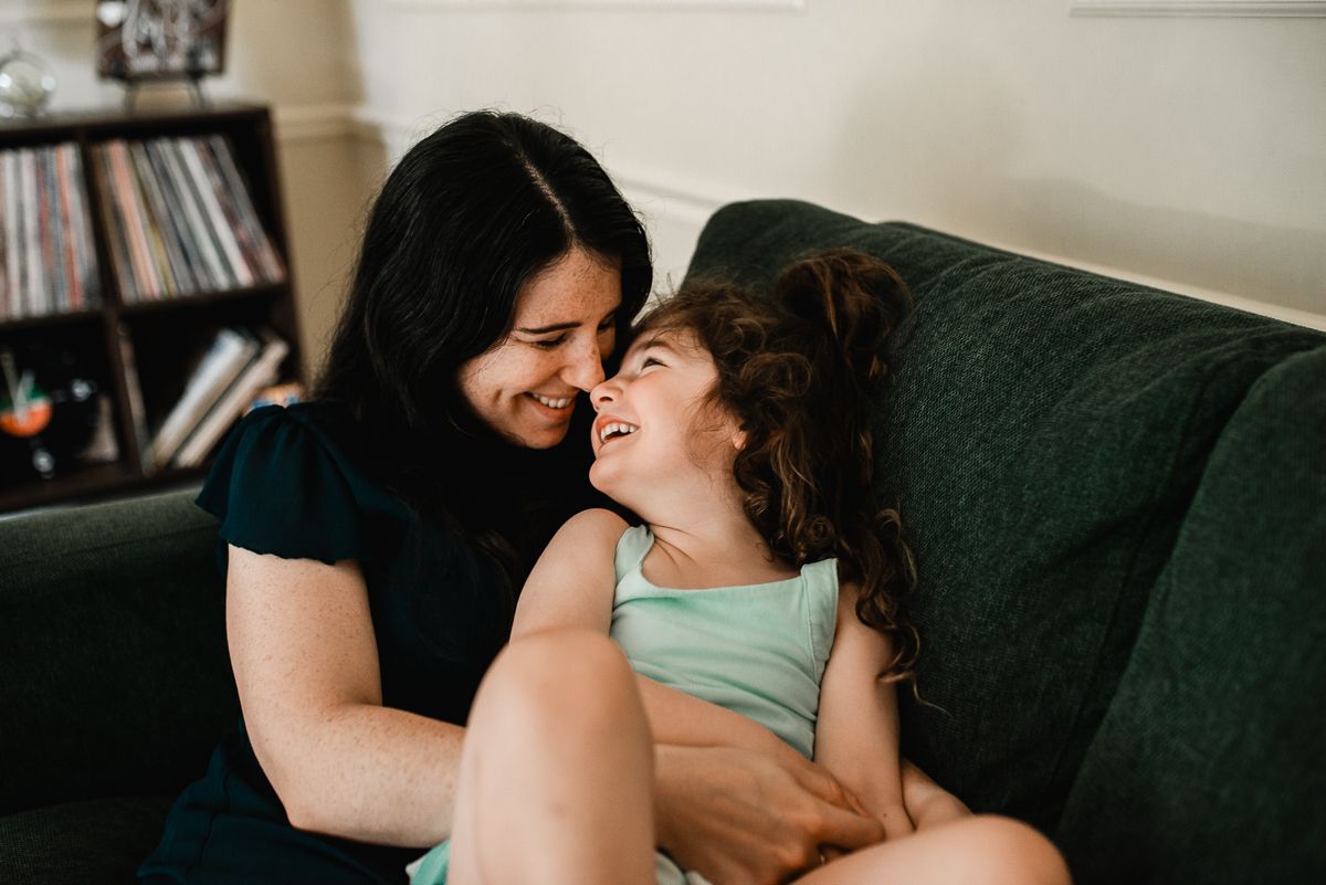 A mother and young daughter share a joyful nose-to-nose moment while sitting together on a green sofa at home.