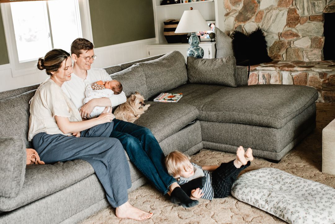 Family of four with newborn and toddler relaxing on gray sectional sofa in cozy living room with stone fireplace.